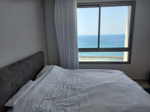 A bedroom with a bed, a window with a view of the sea, and a curtain. - Sea Tower Almog (Haifa)