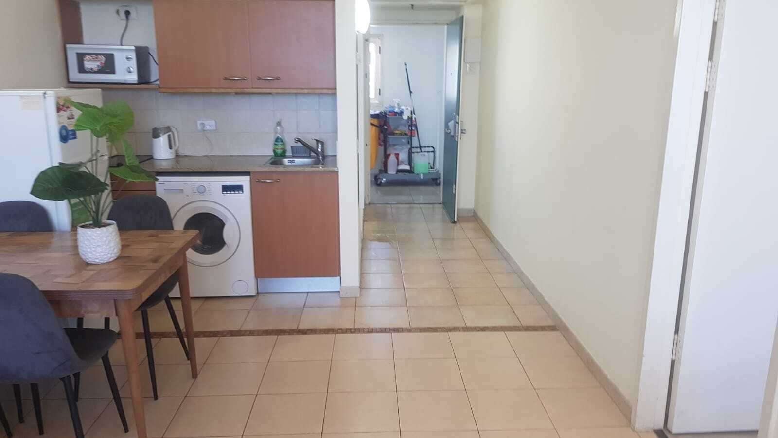 A compact kitchen with a microwave, sink, and washing machine. A dining table with chairs is positioned in front of the kitchen.