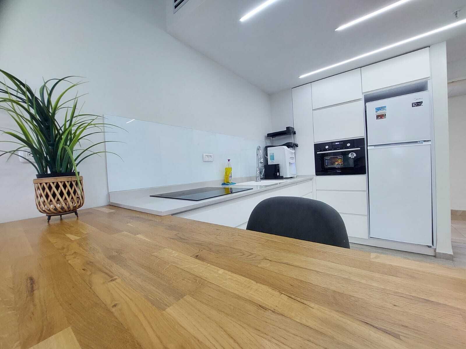 A modern kitchen with white cabinets, a built-in oven, and a refrigerator. There is a wooden dining table with a potted plant on it.