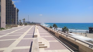 A coastal promenade with a clear view of the sea, a tiled walkway, and a high-rise building on the left.
