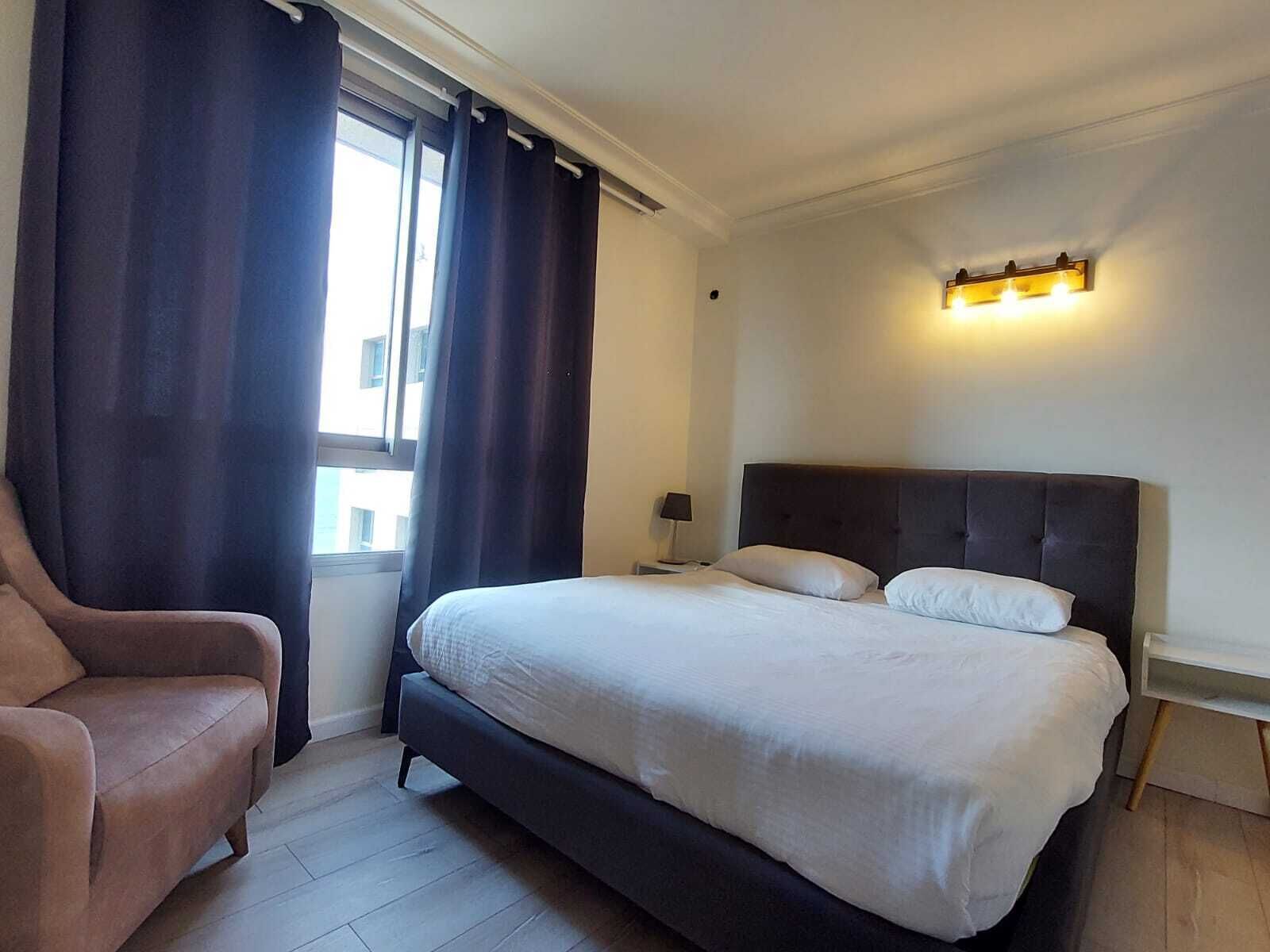A hotel room with a bed, a chair, a nightstand, and a window with curtains.