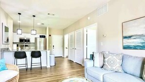 A modern living room with a sofa, coffee table, and a dining area with chairs. There is a kitchen in the background with appliances and a refrigerator. - 2 miles from campus and the square.Cozy 2 BR/2BA in popular Rowendale community (Oxford)
