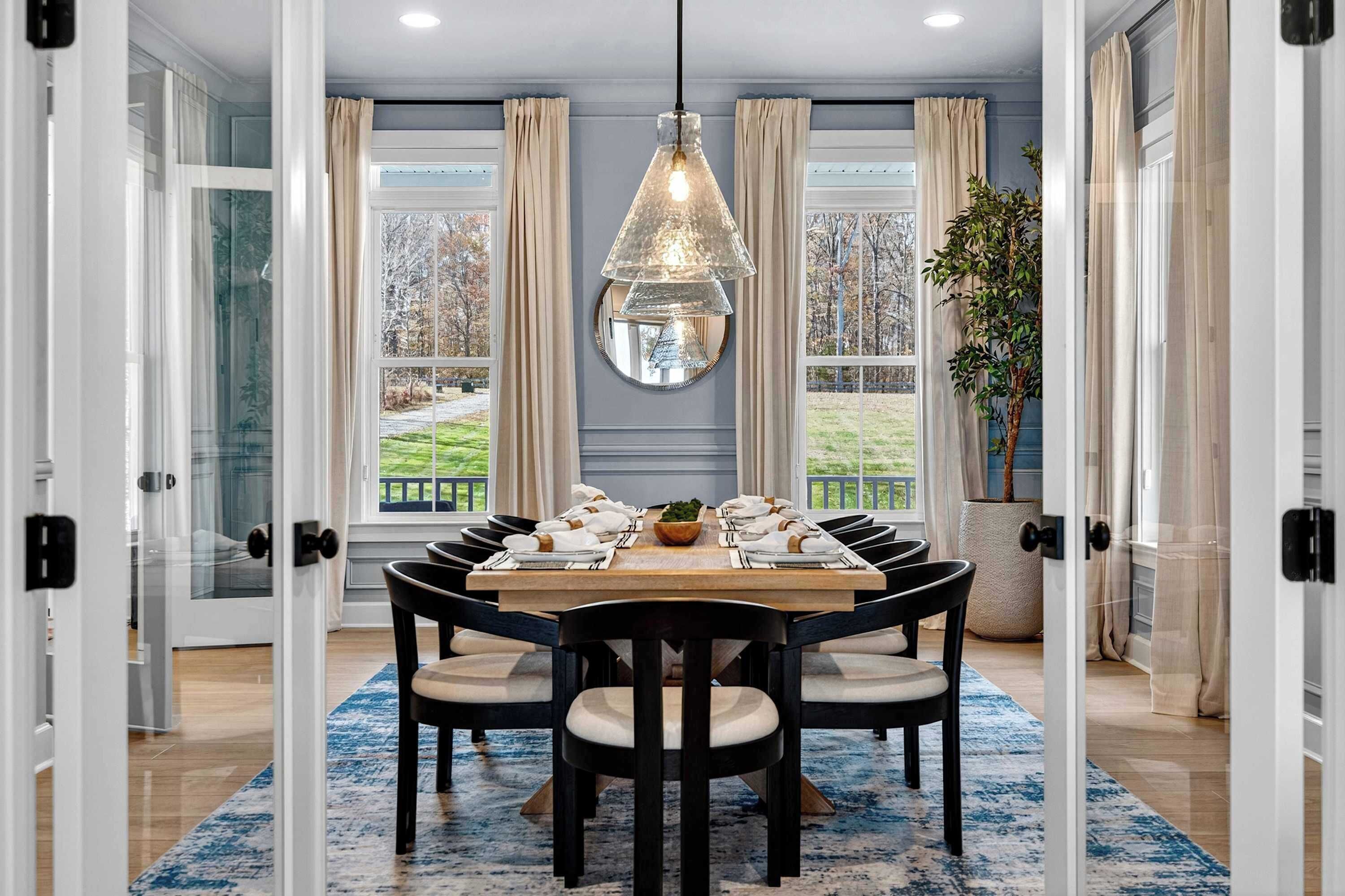 A dining room with a round table, black chairs, a hanging light fixture, and large windows with curtains.