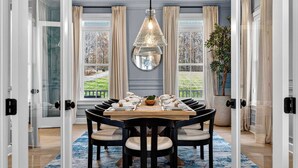 A dining room with a round table, black chairs, a hanging light fixture, and large windows with curtains. - Lakefront Lake Anna - Decks, Views, Hot Tub (Louisa County)