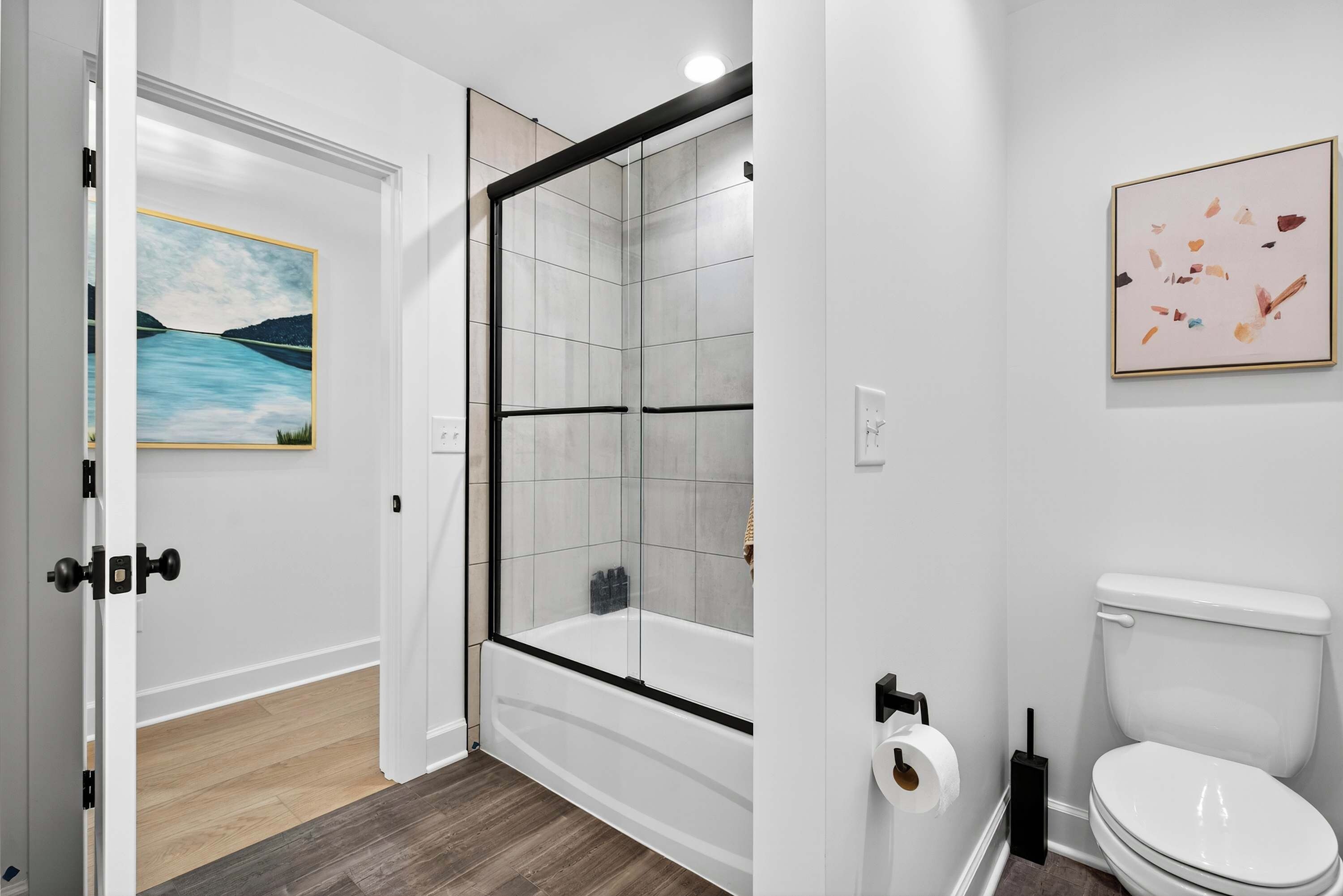 Shared Bath - A tub and shower combo with black-framed glass doors and gray tile, plus a lake painting in the hallway.