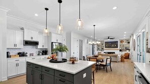 A modern kitchen with white cabinets, a central island, pendant lighting, and a dining area with a table and chairs. - Lakefront Lake Anna - Decks, Views, Hot Tub (Louisa County)