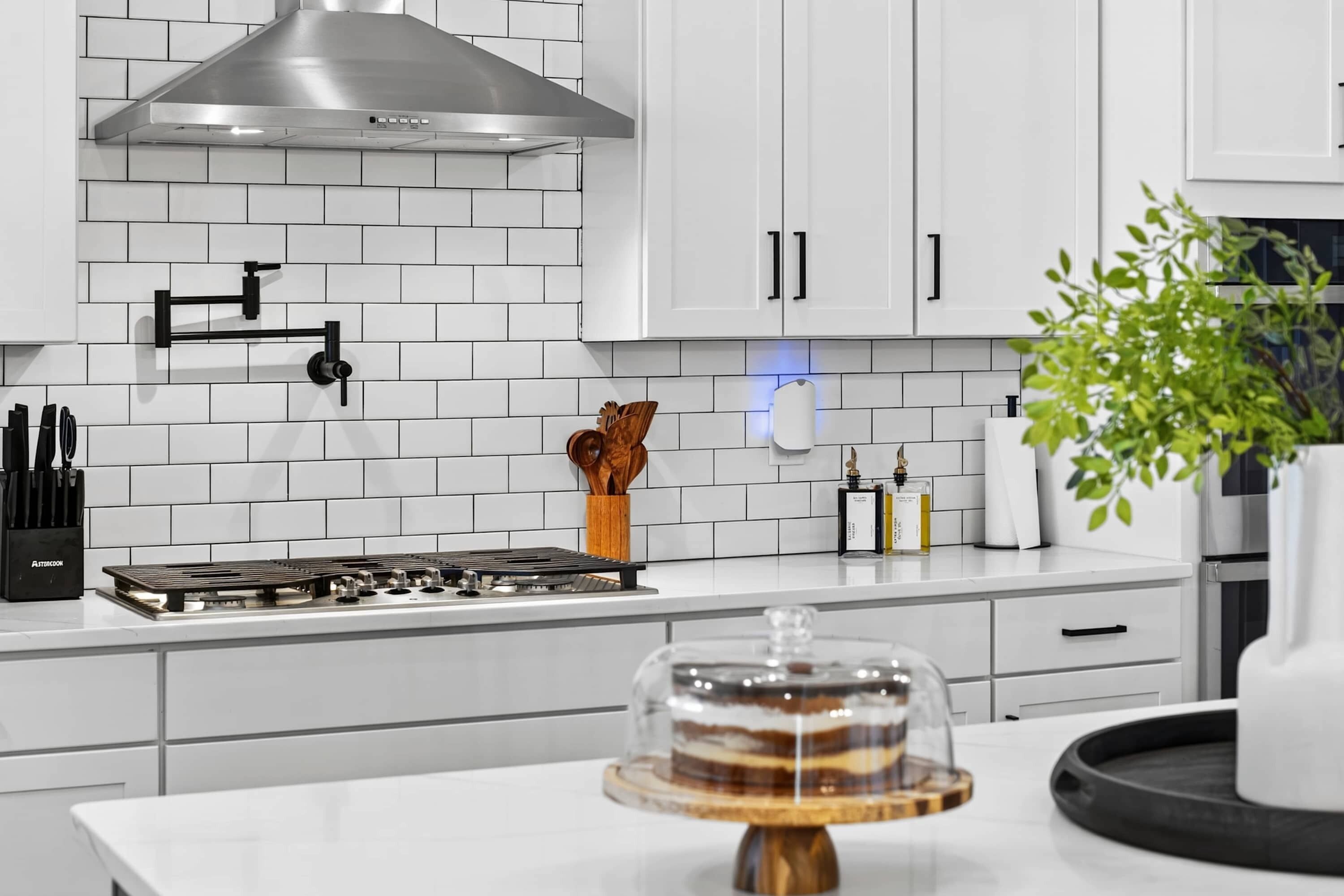 Een moderne keuken met witte kastjes, een roestvrijstalen afzuigkap, een gasfornuis en een taart op een houten standaard.
