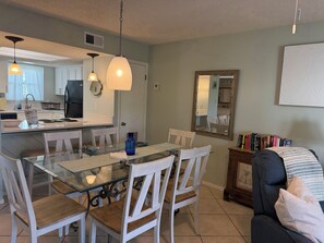 A dining area with a glass table, white chairs, and pendant lights. A kitchen is visible in the background with a refrigerator and a sink. - Cozy 2-bedroom 4 bed condo in Orange Beach directly across the street from Beach (Orange Beach)