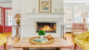 Ein Wohnzimmer mit Kamin, einem Couchtisch mit Büchern und einer Pflanze, einem gemusterten Sessel und einem gelben Sofa.
