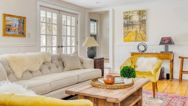 Ein helles Wohnzimmer mit einem beige gefärbten Sofa, einem gelben Sessel, einem Holztisch und einem großen Fenster.