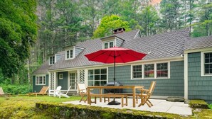 Ein Haus mit einer Terrasse, ausgestattet mit einem Holztisch, Stühlen, einem roten Sonnenschirm und einer Steinmauer.