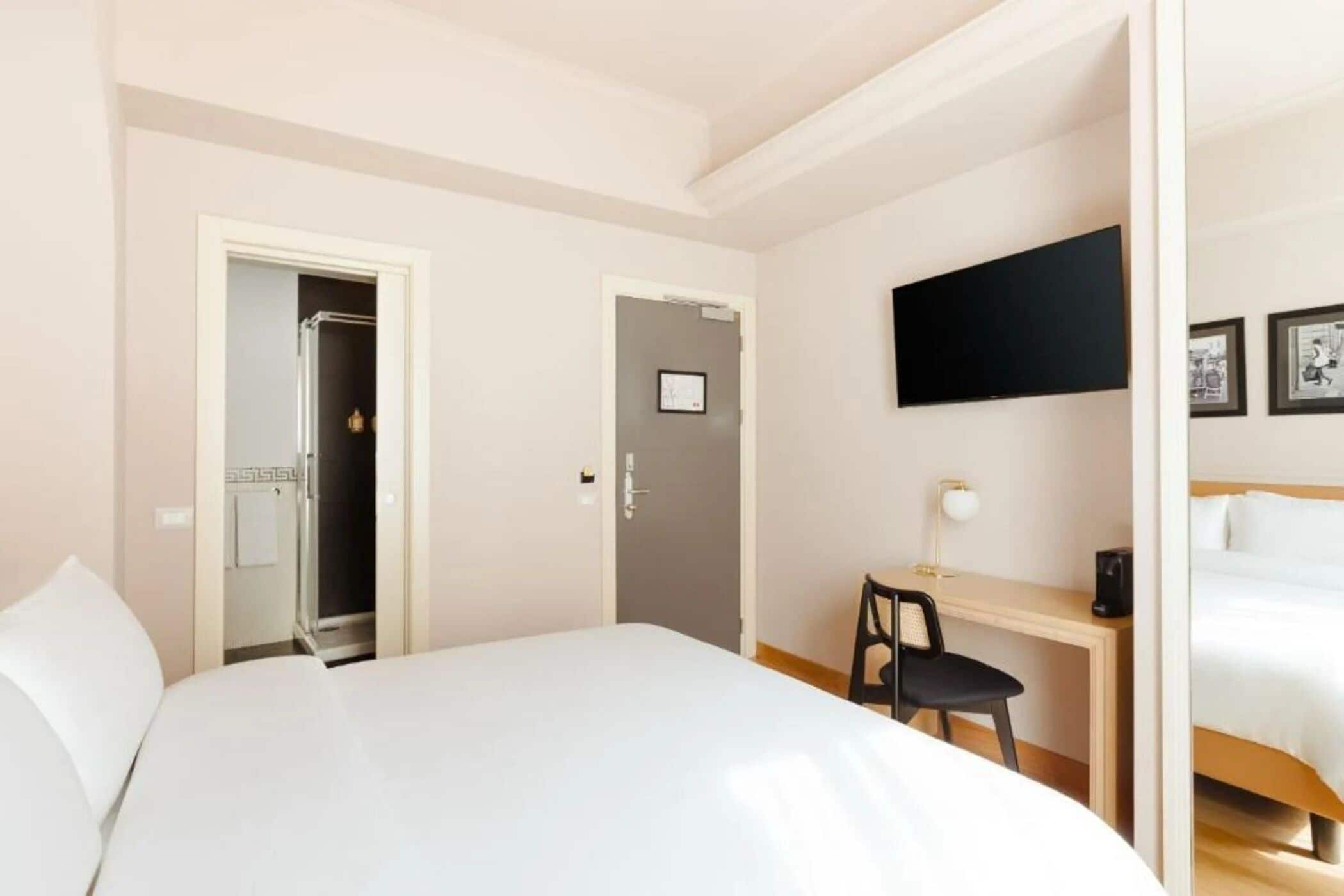 A hotel room with a large bed, a desk with a chair, a TV mounted on the wall, and a door leading to a bathroom.