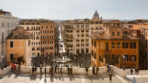 A wide street with buildings on both sides, a central open space, and a dome structure in the distance. - The Couper Piazza di Spagna (Rome)