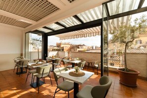 An outdoor dining area with multiple tables and chairs, large windows, and a view of the cityscape. - The Couper Piazza di Spagna (Rome)