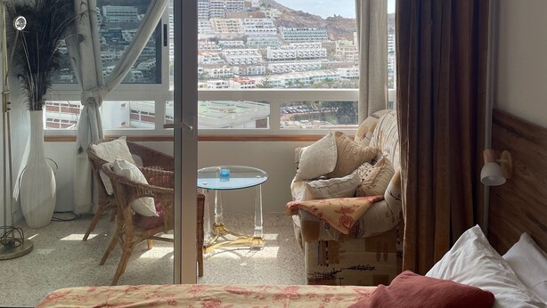 Habitación de hotel con cama, una silla, una mesita y vistas a un paisaje urbano a través de un amplio ventanal.