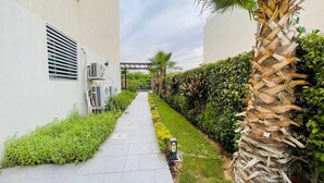 A narrow pathway flanked by lush greenery and a palm tree, leading to a building with air conditioning units. - فيلا للايجار اليومي خمس غرف داخل كمبوند بالم هيلز
villa for daily rent (Giza Governorate)