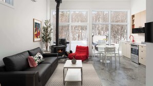 Un salon moderne avec un canapé en cuir noir, une table basse blanche et un fauteuil rouge. On aperçoit une cheminée au fond, et il y a un coin cuisine avec des meubles blancs et un fourneau noir.