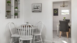 Un coin repas avec des chaises blanches, une table ronde et une chaise gris foncé. Des plantes en pot sont disposées sur des étagères et un tableau encadré orne le mur.