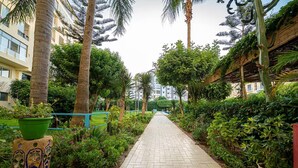 Een pad omzoomd met palmbomen en diverse planten, dat leidt naar een gebouw met een blauwe balustrade.