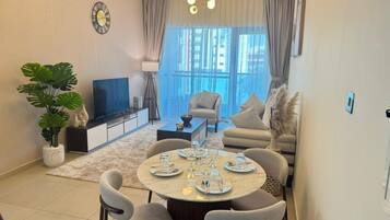 A modern living room with a round dining table set for four, a sofa, a TV, and a large window with curtains.