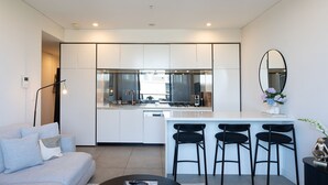 A modern kitchen with white cabinets, a central island with bar stools, a large mirror, and a dining area with a sofa and coffee table. - Riverside Parramatta I90 (Parramatta)