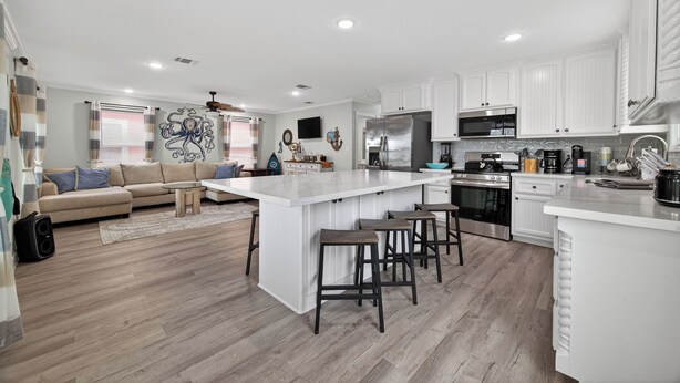 A modern kitchen with white cabinetry, a large island with stools, and a dining area with a sofa and coffee table. - Pirate's Bay. New build! Pets! Hot Tub! Affordable! Views of Ocean AND Bay! (Surfside Beach)