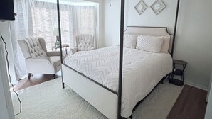 Una cama con dosel con cubrecama blanco, un ventilador de techo y vistas a una ventana con cortinas traslúcidas.