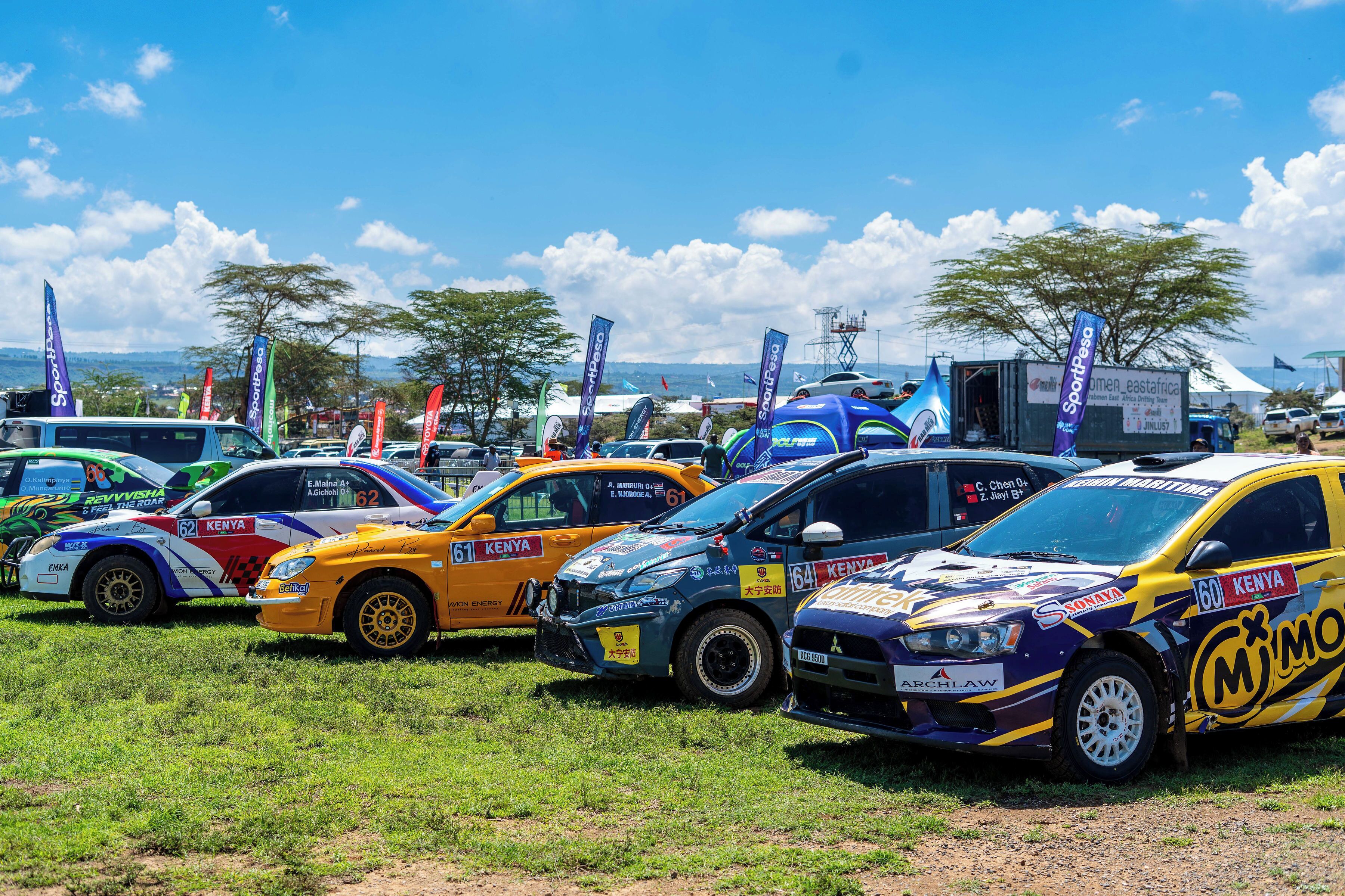 A row of rally cars parked in a grassy field with flags and tents in the background.