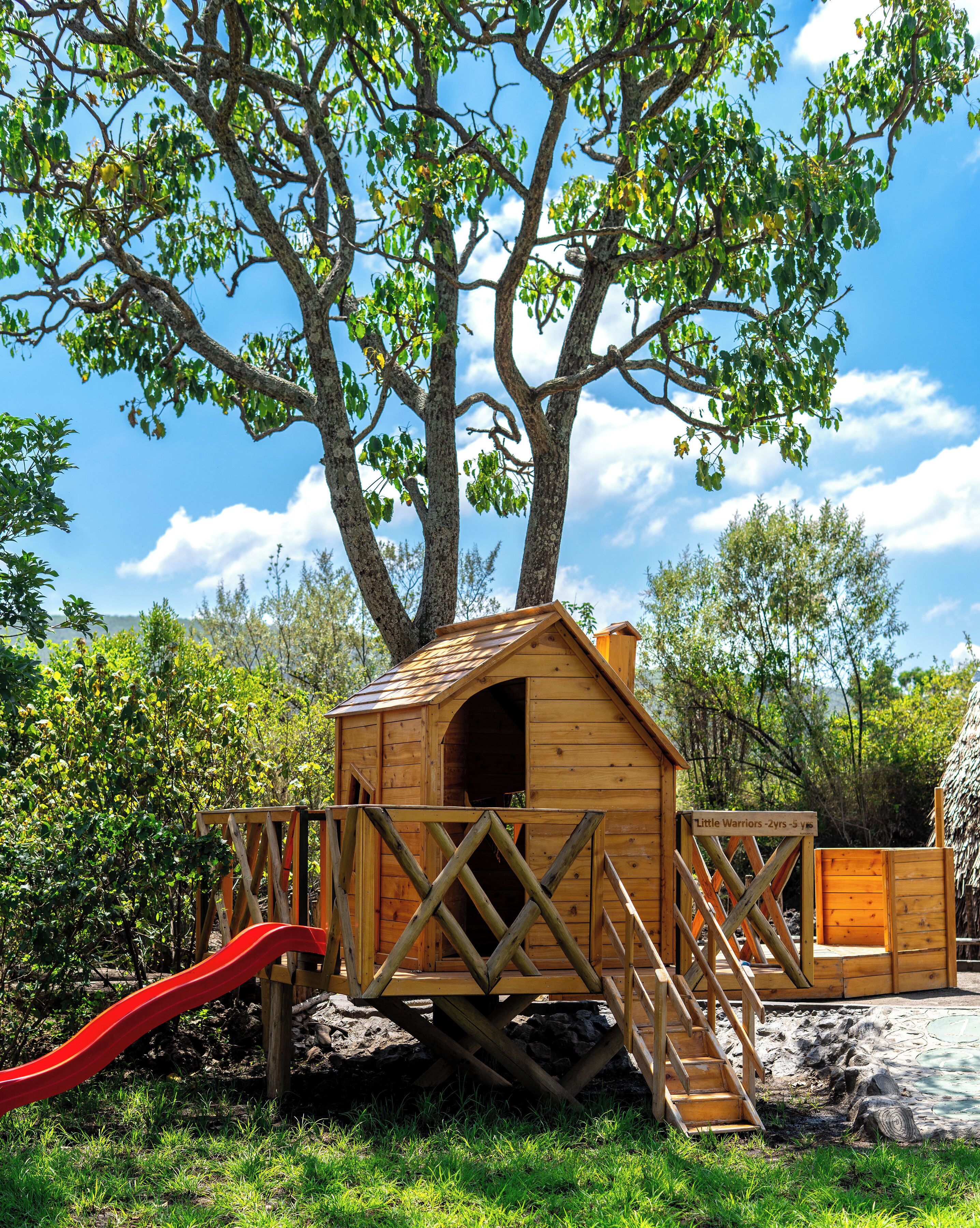 A wooden playhouse with a slide, surrounded by trees and a clear sky.