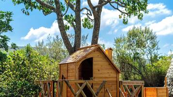 A wooden playhouse with a slide, surrounded by trees and a clear sky.