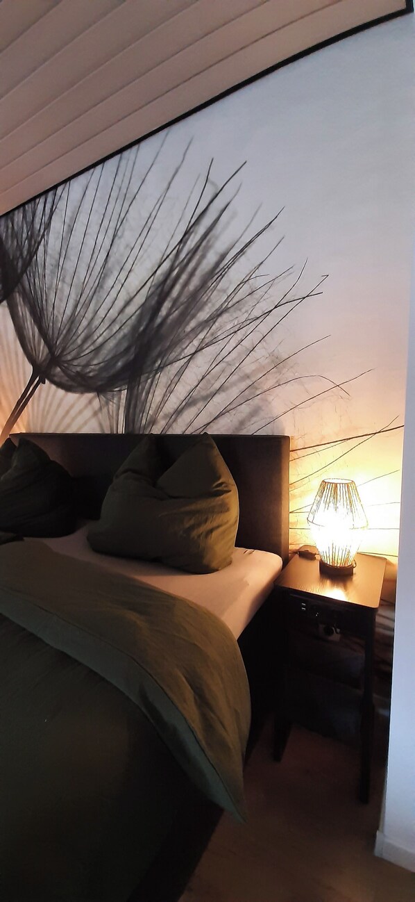 Un dormitorio con cama, almohadas, una mesita de noche con lámpara y una pared con diseño de hojas.
