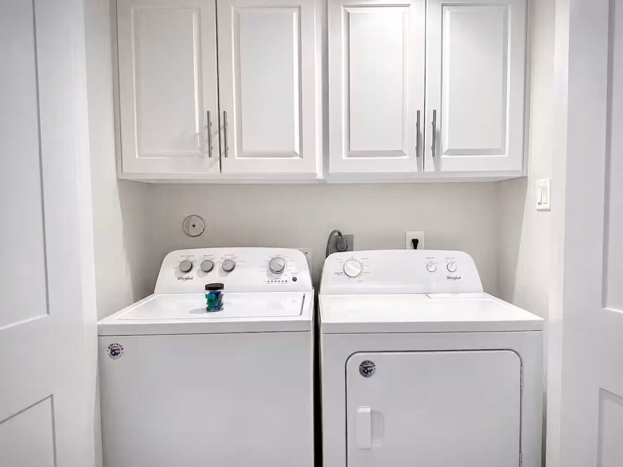 A laundry room with two white front-loading washers and dryers, white cabinets above, and a small blue container on the left washer.