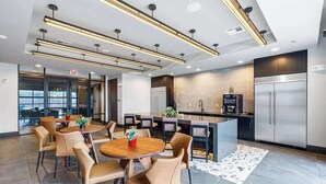 Un espace de repas moderne avec une table ronde centrale et des chaises, une cuisine équipée d’appareils électroménagers en acier inoxydable et une vue sur la ville grâce à de grandes fenêtres.