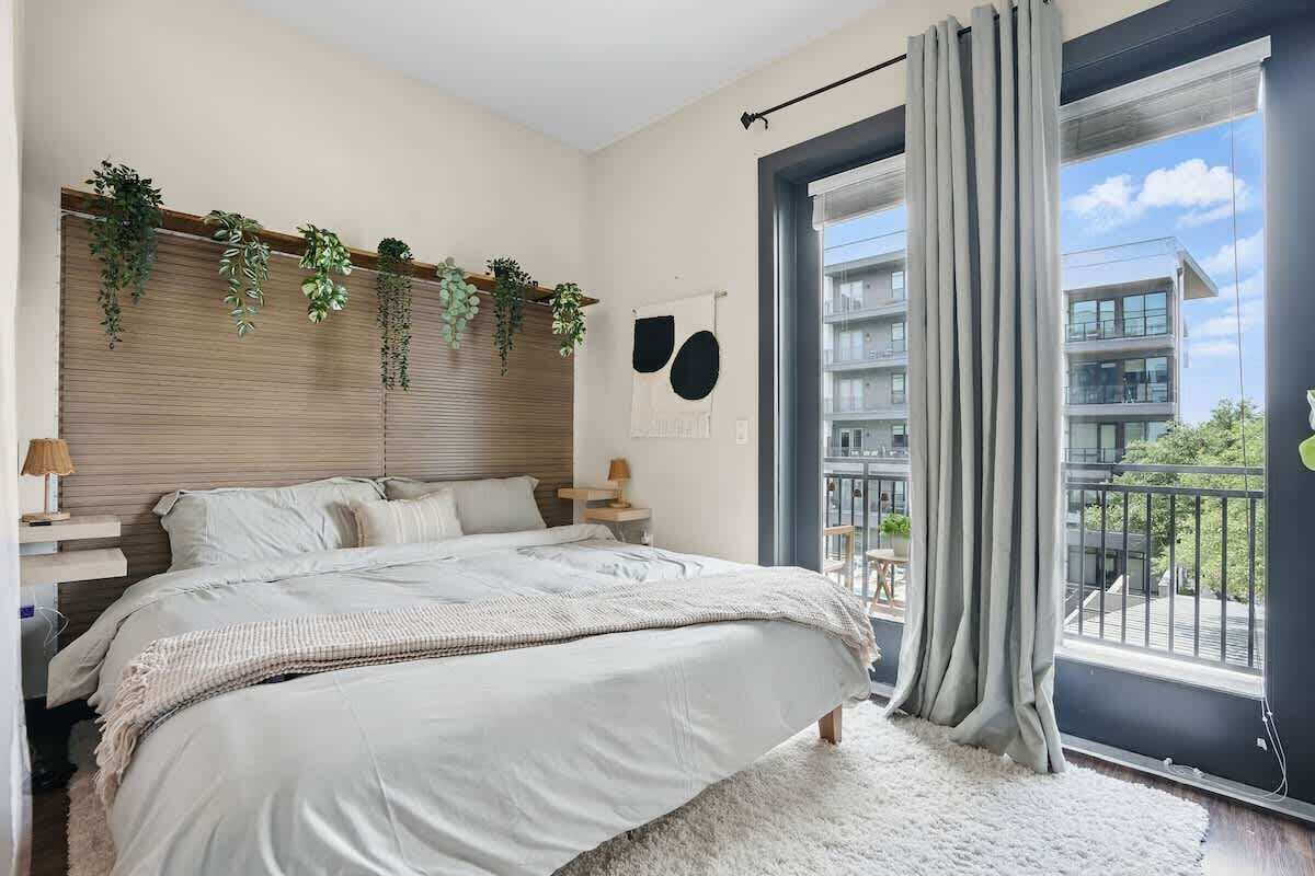 Ein Schlafzimmer mit einem großen Bett, einem Fenster mit Vorhängen, einem Balkon und Blick auf Gebäude.