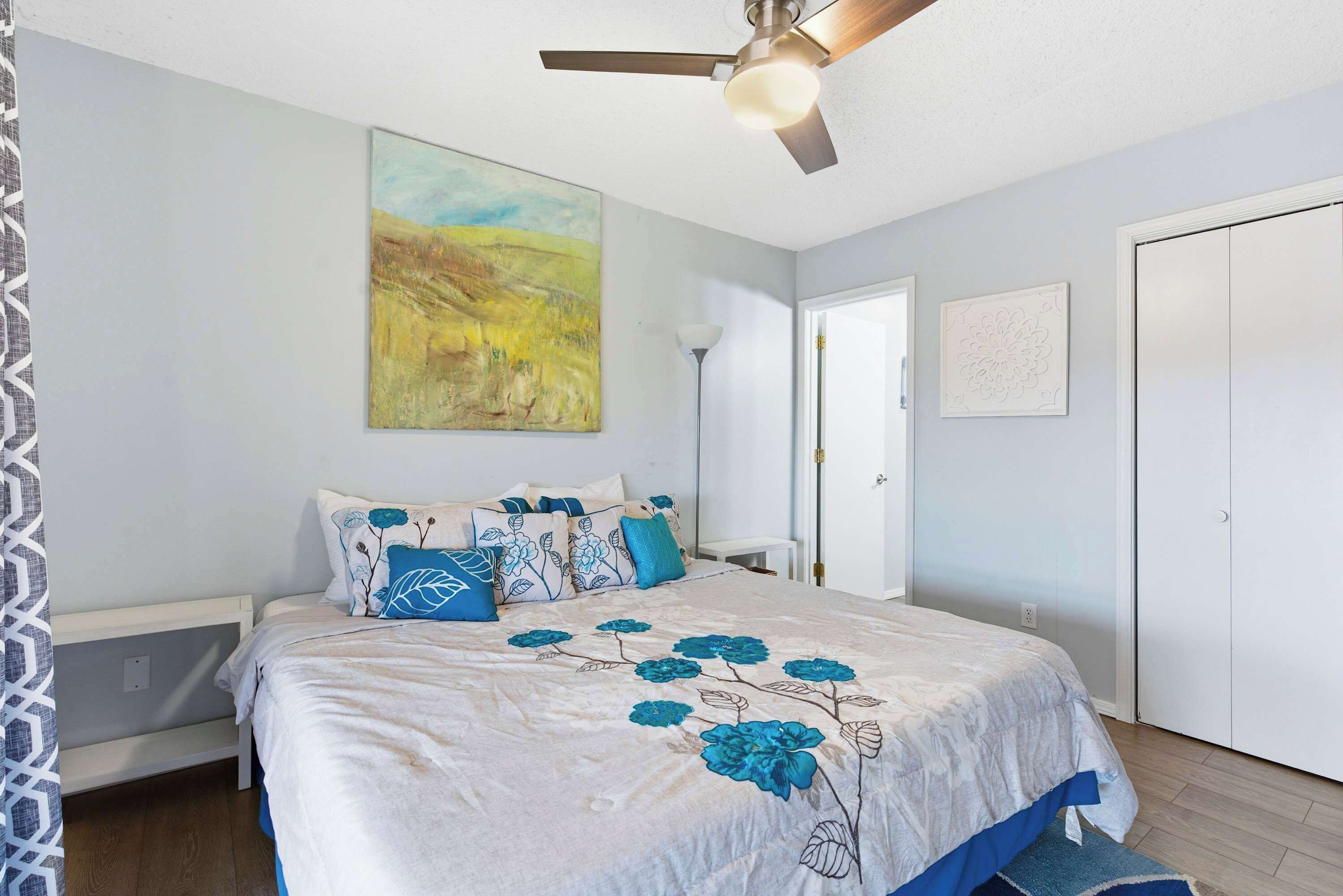 A bedroom with a bed, a ceiling fan, a painting, and a door.