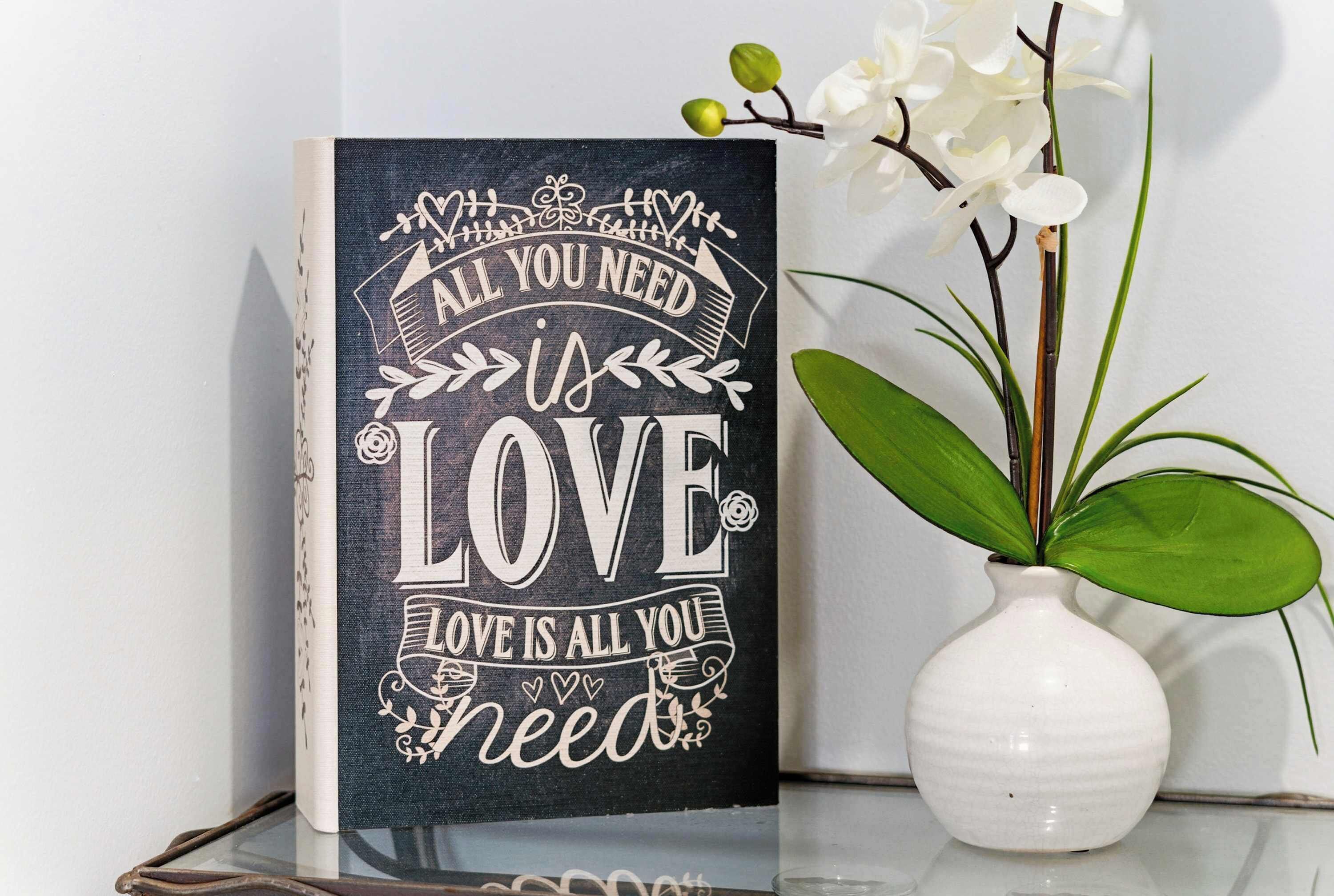 A decorative sign with the phrase "All you need is love" placed next to a white vase with a plant.