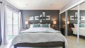 A modern bedroom with a large bed, two bedside tables with lamps, a sliding glass door, and cityscape artwork on the wall. - Cozy 2-bedroom condo in peaceful studio city with AC (Burbank)