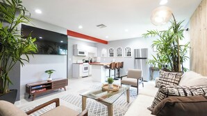 A modern living room with a glass coffee table, a sofa, and a TV. There is a kitchen in the background with white cabinets and a stainless steel refrigerator. - Cozy 2-bedroom condo in peaceful studio city with AC (Burbank)