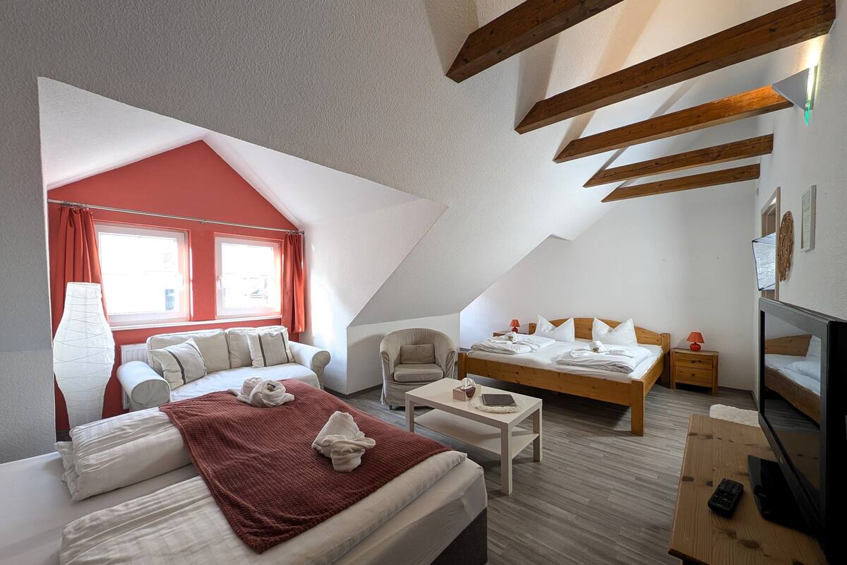 Una habitació d'hotel moderna amb un sostre inclinat, un llit gran amb un llençol vermell, un escriptori amb un ordinador i una televisió.