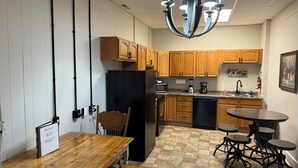 Cocina compacta con armarios de madera, un refrigerador negro, microondas, cafetera y un lavamanos. Hay una mesa de comedor de madera con bancos y una araña colgante.