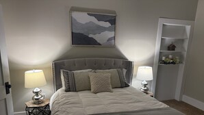 Un dormitorio con cabecera capitonada, cama con almohadas estampadas, dos lámparas de noche, un cuadro abstracto enmarcado sobre la cama y una estantería blanca con objetos decorativos.