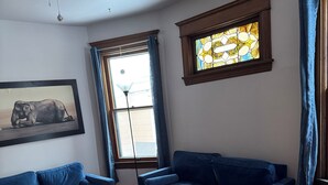 Un salon avec deux canapés bleus, un tapis à motifs, un ventilateur de plafond, un vitrail et un tableau représentant un éléphant, encadré.
