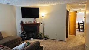 A living room with a fireplace, a television mounted on the wall, and a dining area visible through an open door. - 3-bedroom house in lancaster. (Lancaster)