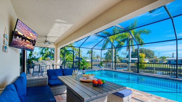 A poolside area with a dining table, blue sofas, and a TV mounted on the wall. - Villa Sunny Dream - New Villa - perfect for families and boaters (Cape Coral)
