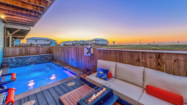 A pool area with a wooden deck, a fire feature, and a sofa with cushions.