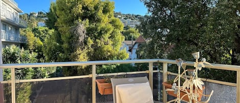 Ein Balkon mit einem Metalltisch und -stühlen, einem Topfpflanzen und Blick auf einen Baum und Gebäude.