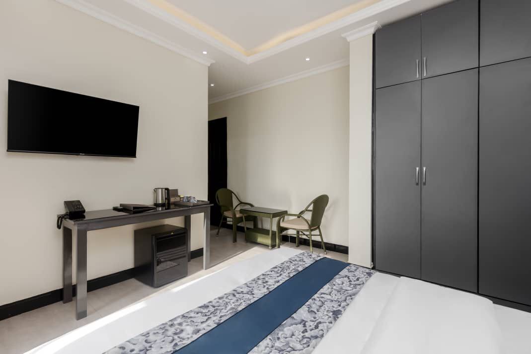 A modern hotel room with a bed, a desk with a phone and a coffee maker, a TV mounted on the wall, and a wardrobe.