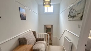A narrow, carpeted hallway with a wooden cabinet, a cushioned bench, a radiator, and a window at the end. - Welcome to The Pink House managed by Sean! (Torquay)