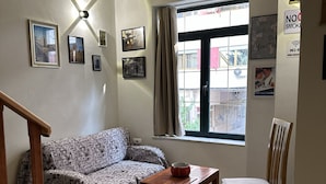 Una sala de estar con un sofá, una mesa de madera con un cuenco, una silla de madera, una escalera y una ventana con cortinas.