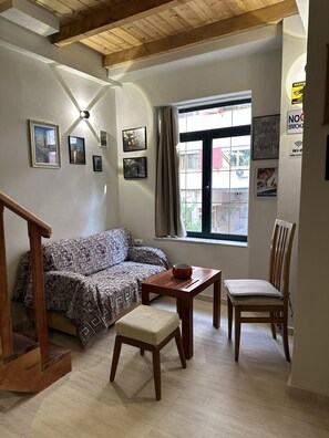 Un salon avec un canapé, une table en bois sur laquelle se trouve un saladier, une chaise en bois, un escalier et une fenêtre avec des rideaux.